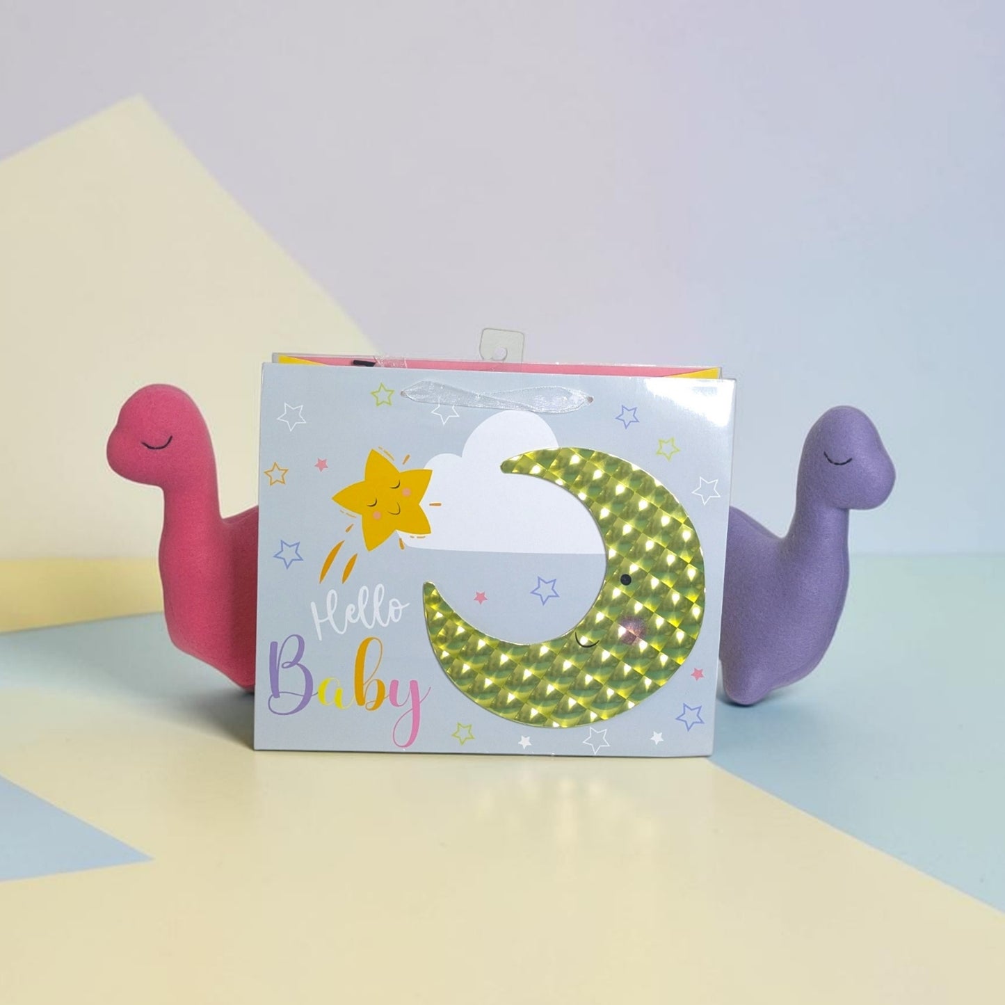 A baby shower favor box with a 'Hello Baby' theme, flanked by two small dinosaur-shaped favors, one pink and one purple, on a pastel geometric background.