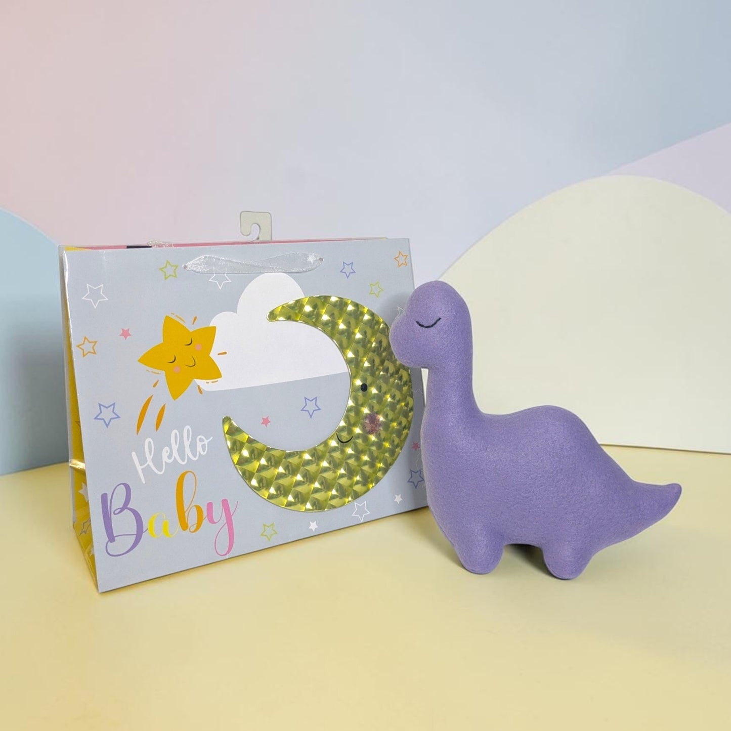 A purple dinosaur-shaped plush toy sits next to its illustrated gift box. The box is light gray with a yellow star, white cloud, and a glittery green crescent moon, plus the words 'Hello Baby' in pastel lettering.