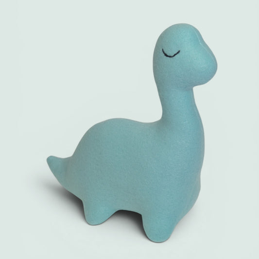 A light blue dinosaur-shaped plush toy with a simple embroidered closed-eye smile, photographed on a clean light gray background.