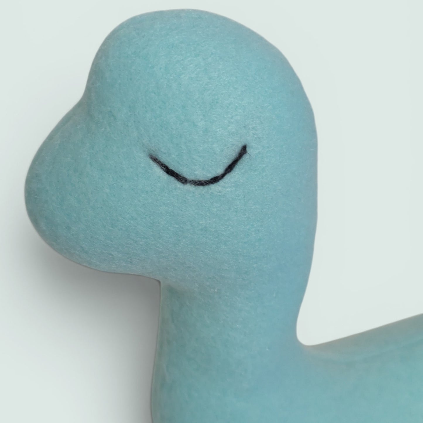 A plush dinosaur toy made of soft blue felt with a black stitched eye, standing against a solid pale blue background.