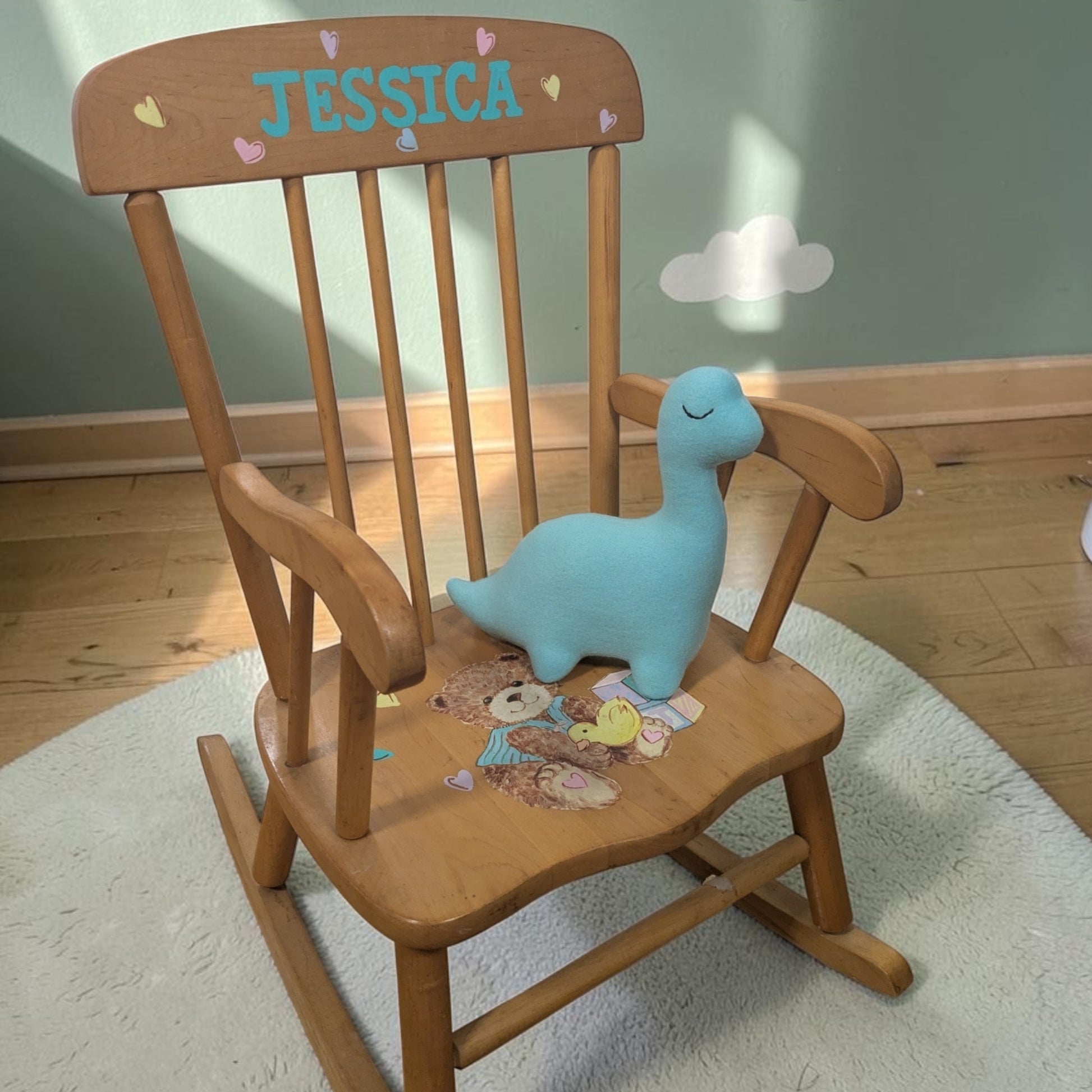 A wooden rocking chair with the name 'JESSICA' and small hearts painted on the backrest, styled with a light blue plush dinosaur toy on the seat in a nursery setting.