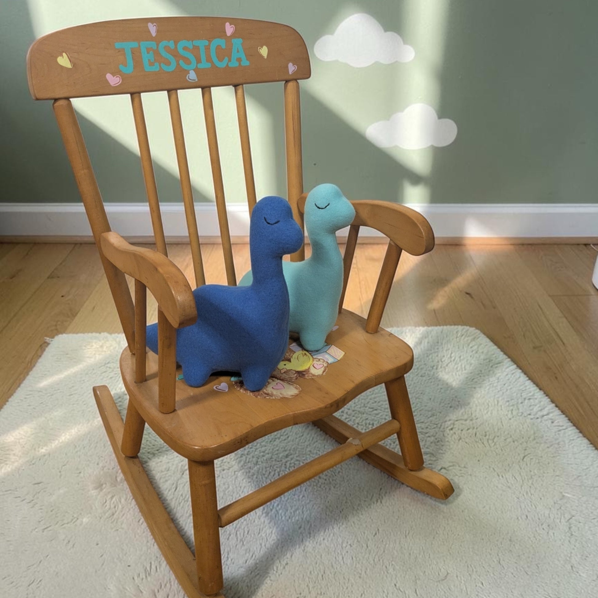 A wooden rocking chair in a child's room, personalized with the name 'JESSICA' on the backrest. Two plush dinosaur toys, one blue and one light blue, are sitting on the chair.