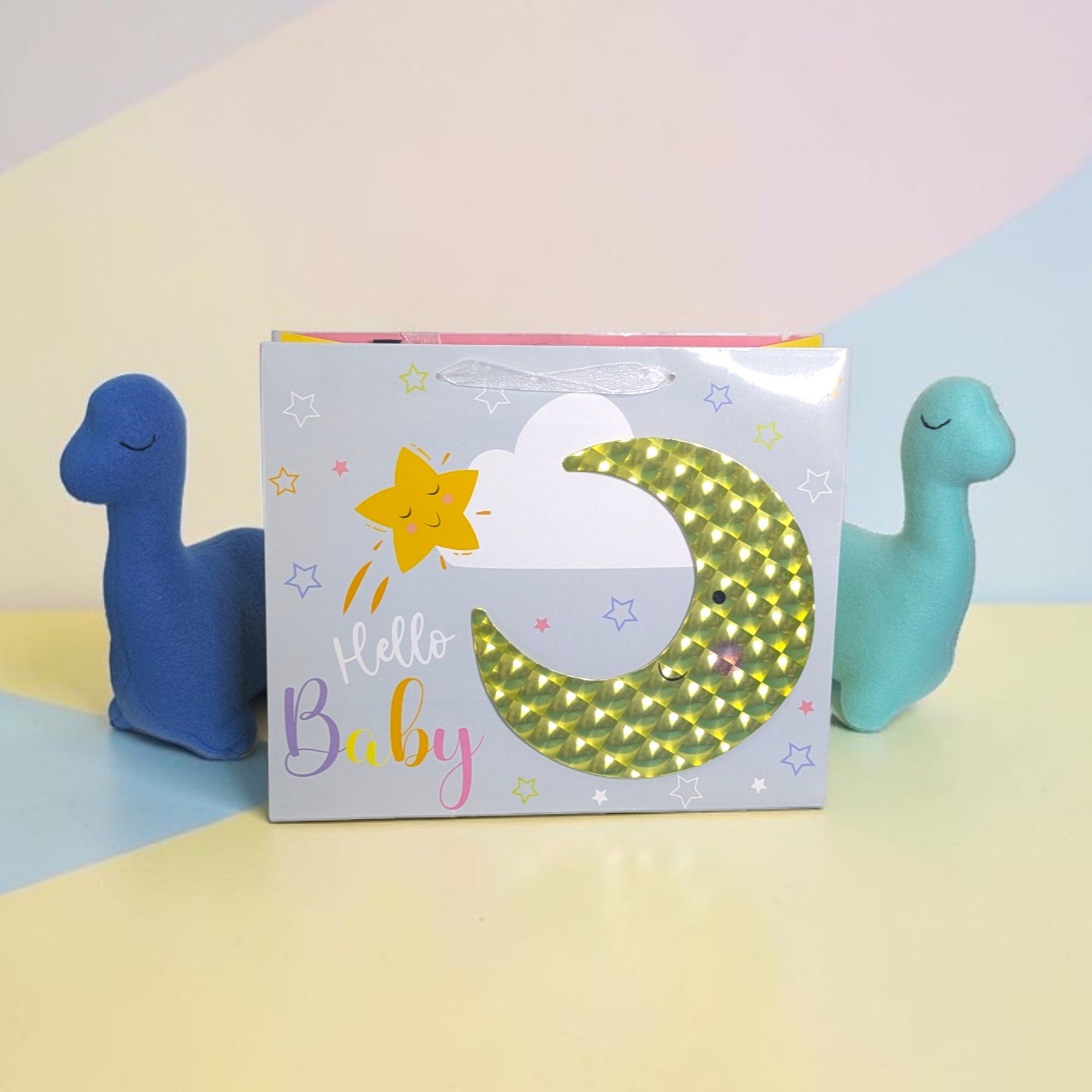 A baby gift box with a holographic gold crescent moon and 'Hello Baby' text, flanked by two plush dinosaur toys in blue and teal on a pastel background.