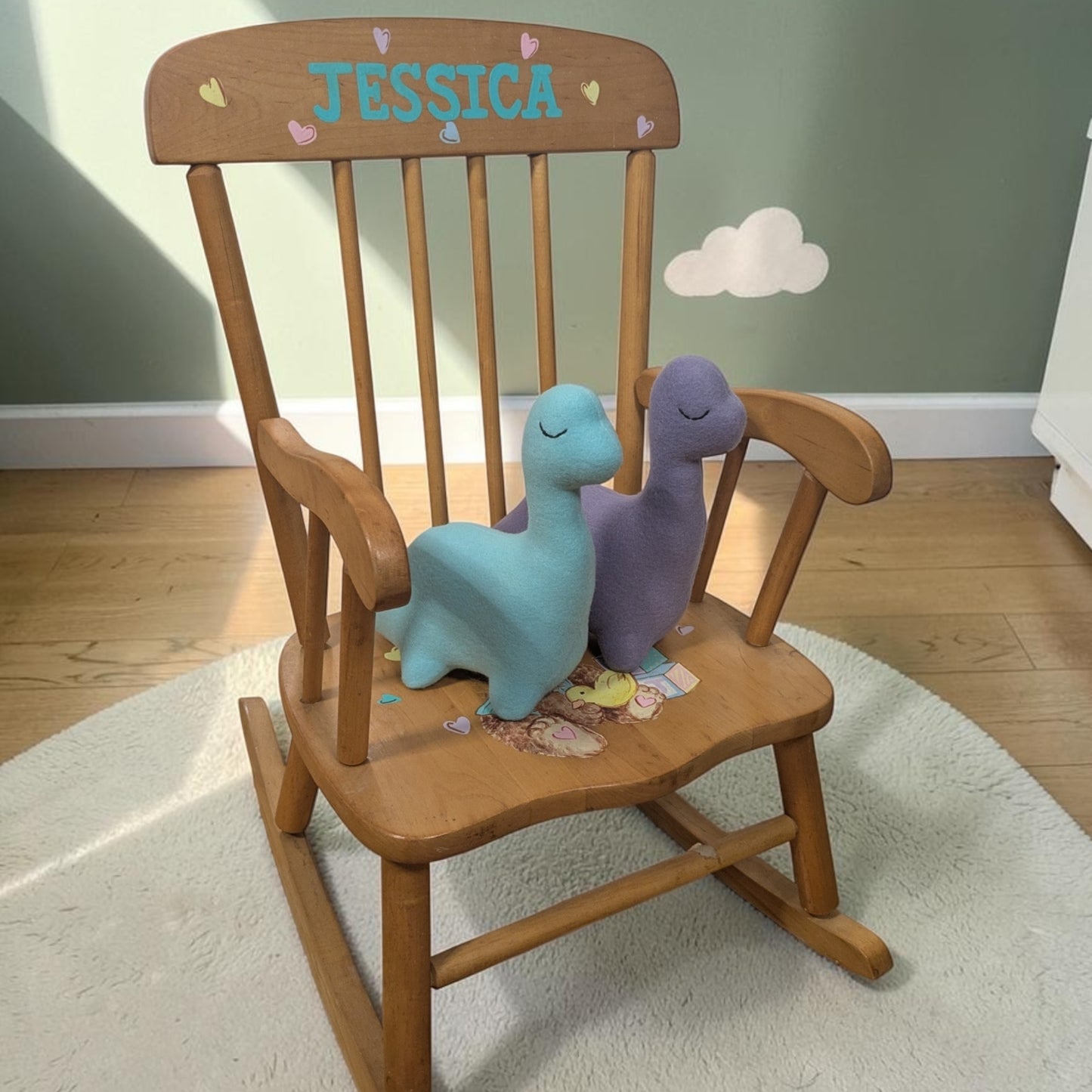 A wooden rocking chair with the name 'JESSICA' and small hearts painted on the backrest, styled with two plush dinosaur toys on a white rug in a child's room.