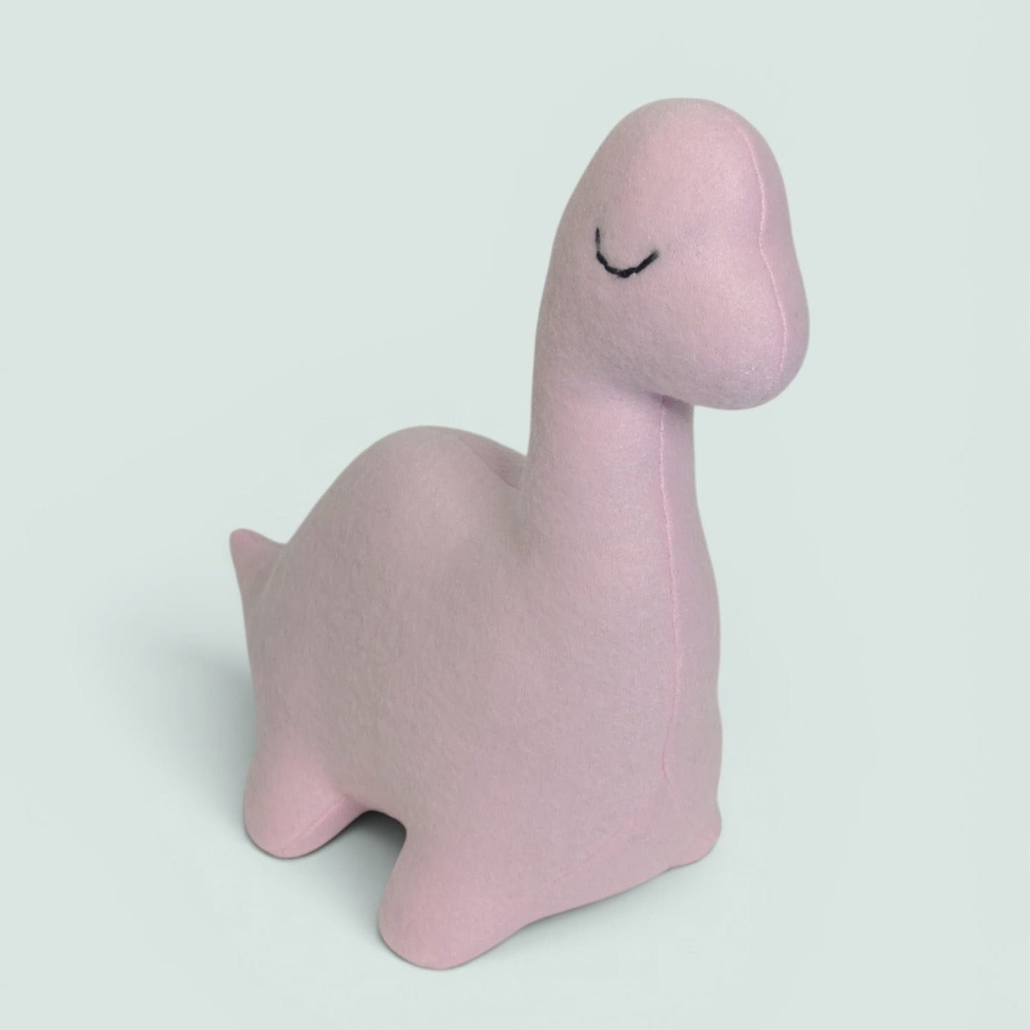 A light pink plush dinosaur toy with a simple black stitched closed-eye detail, shown on a solid pale gray background.
