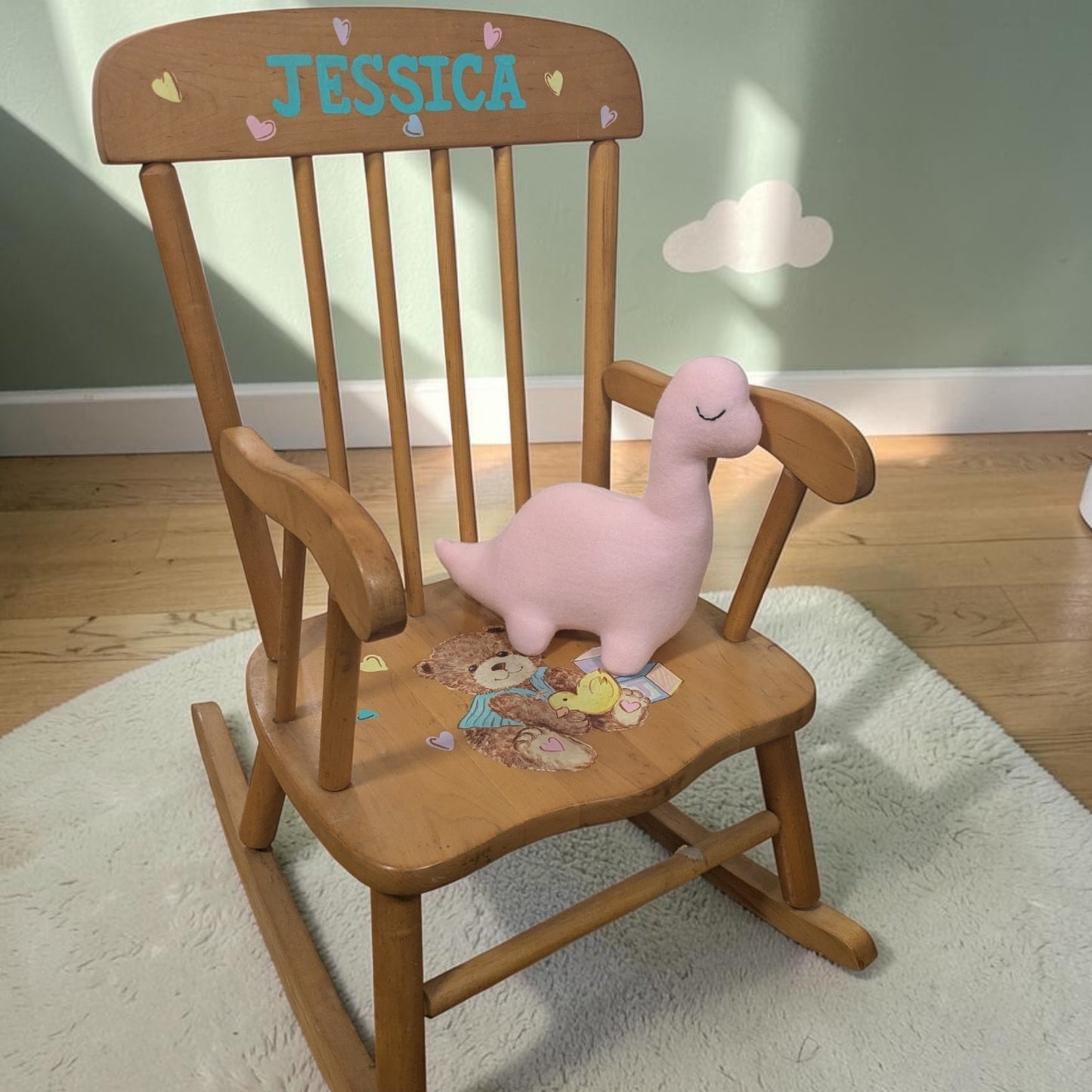 A wooden rocking chair with the name 'JESSICA' painted on the backrest, styled with a pink plush dinosaur toy on the seat, set on a white rug in a child's room.
