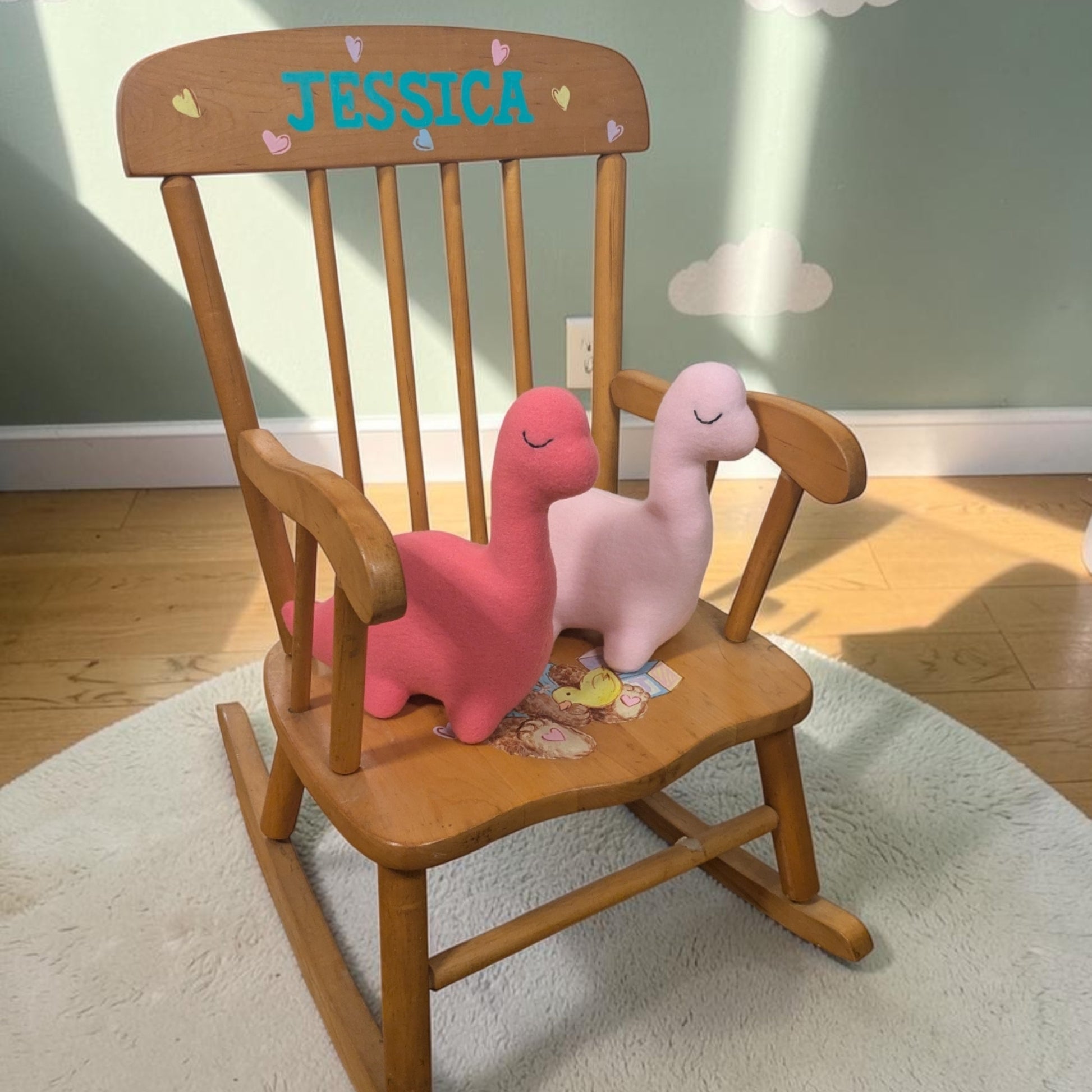 A wooden rocking chair with a personalized backrest reading 'JESSICA' in blue, featuring two plush dinosaur toys on the seat, set on a white rug in a child's room.