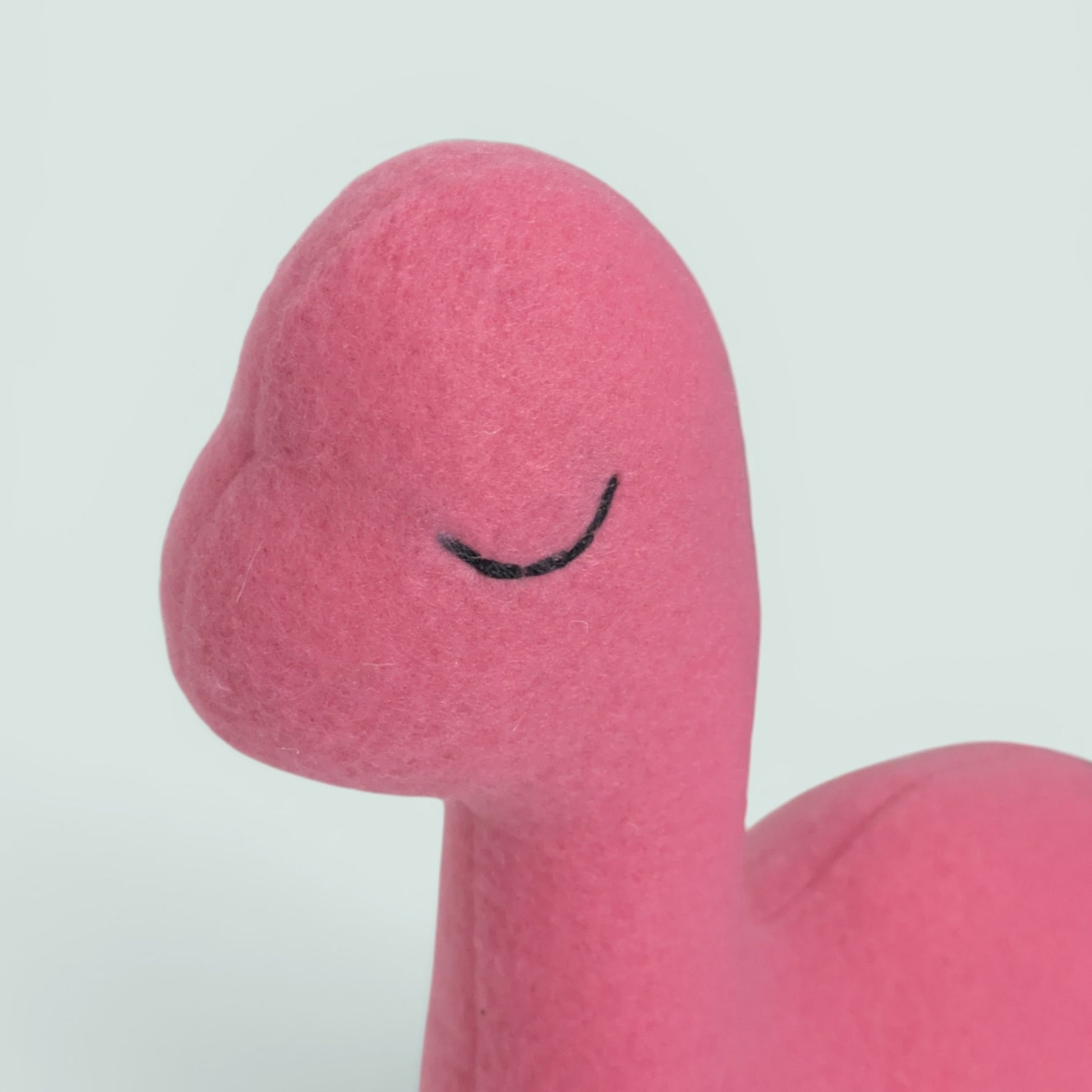 A close-up of a pink plush toy with a simple embroidered black curved line for a closed eye, shown against a plain light gray background.