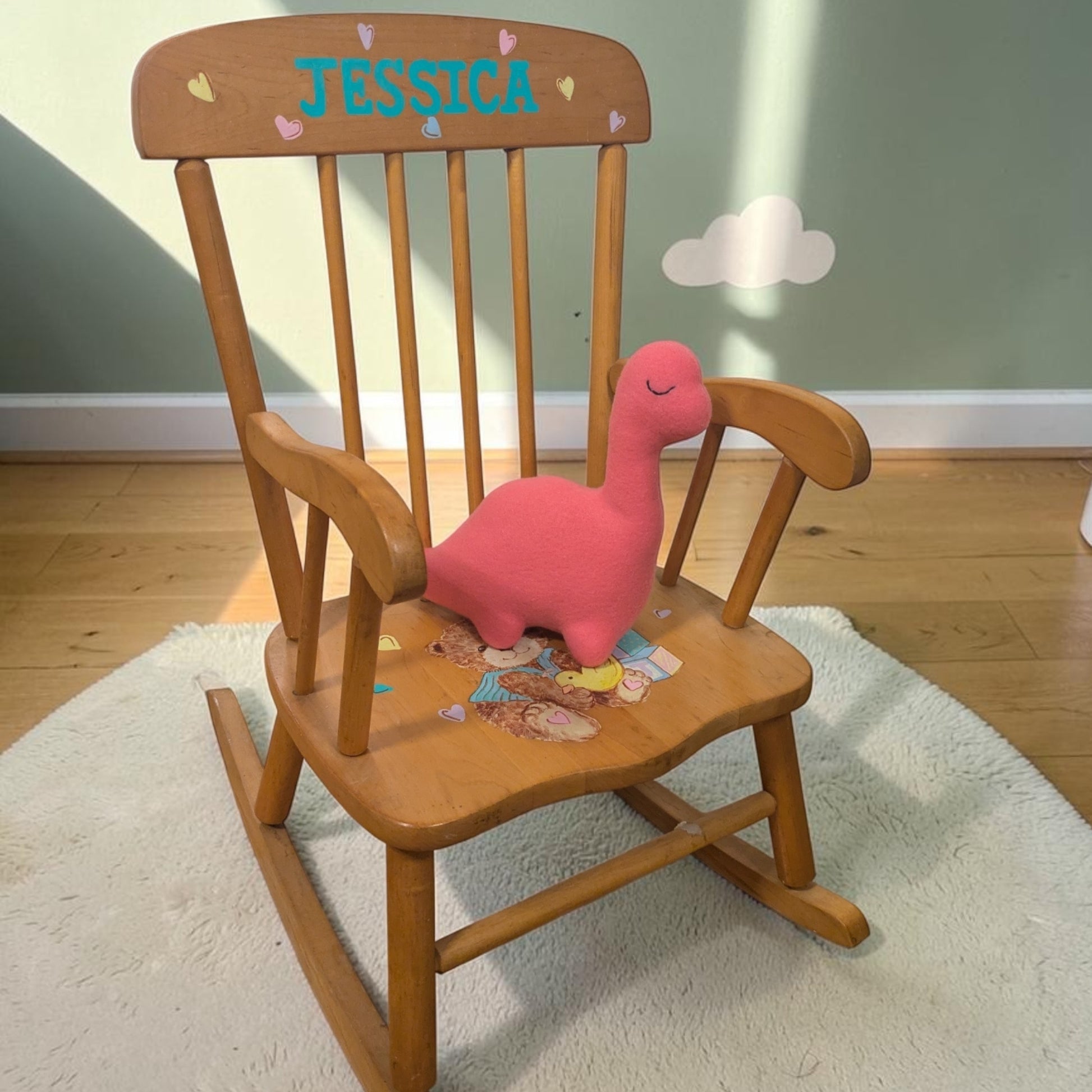 A wooden children's rocking chair with 'JESSICA' painted on the backrest, featuring a pink plush dinosaur toy on the seat, set on a white rug in a room with a green wall and wood floor.