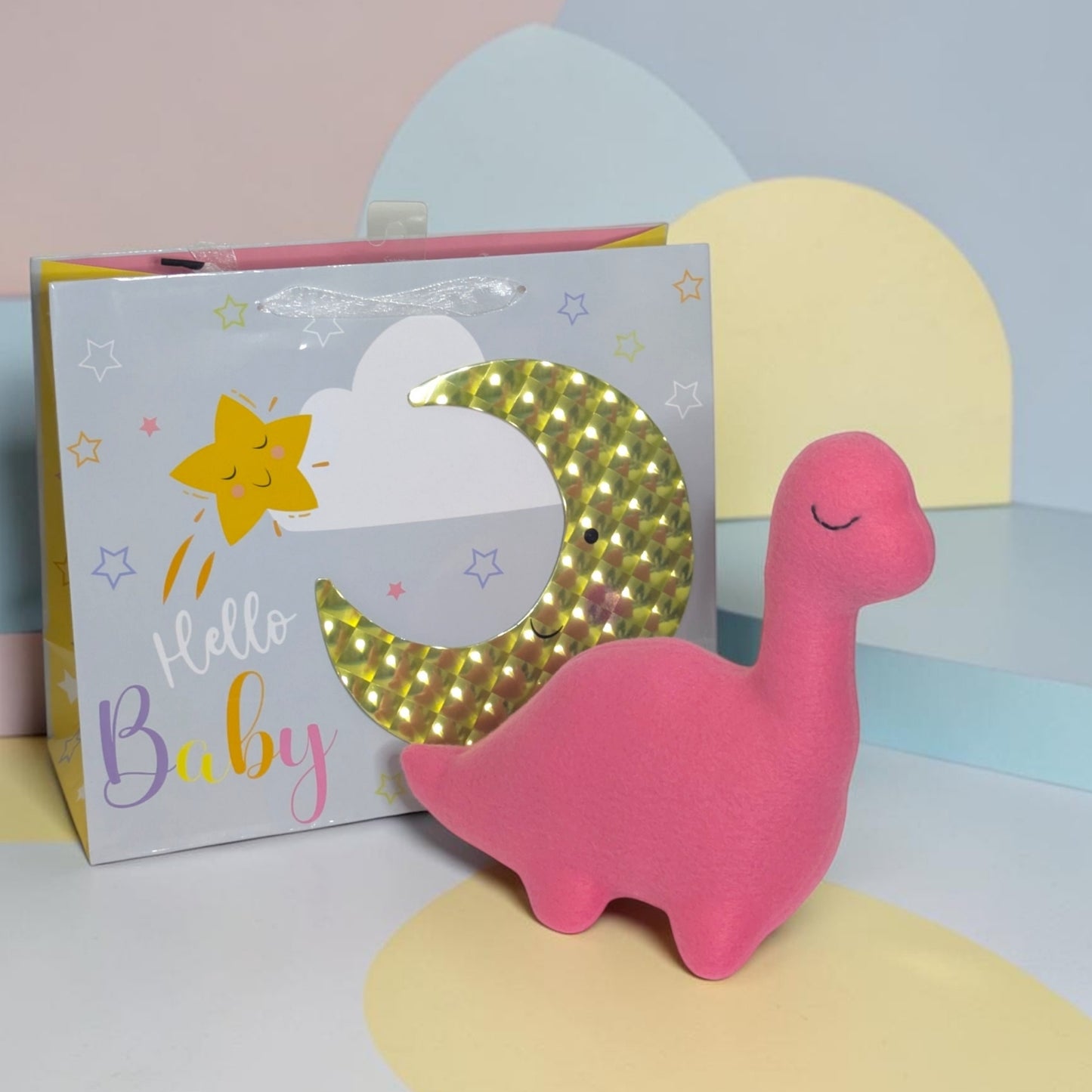 A pink plush dinosaur toy with a sleeping face, posed in front of its gift box. The box is white with pastel lettering reading 'Hello Baby', a yellow star, and a glittery gold crescent moon.
