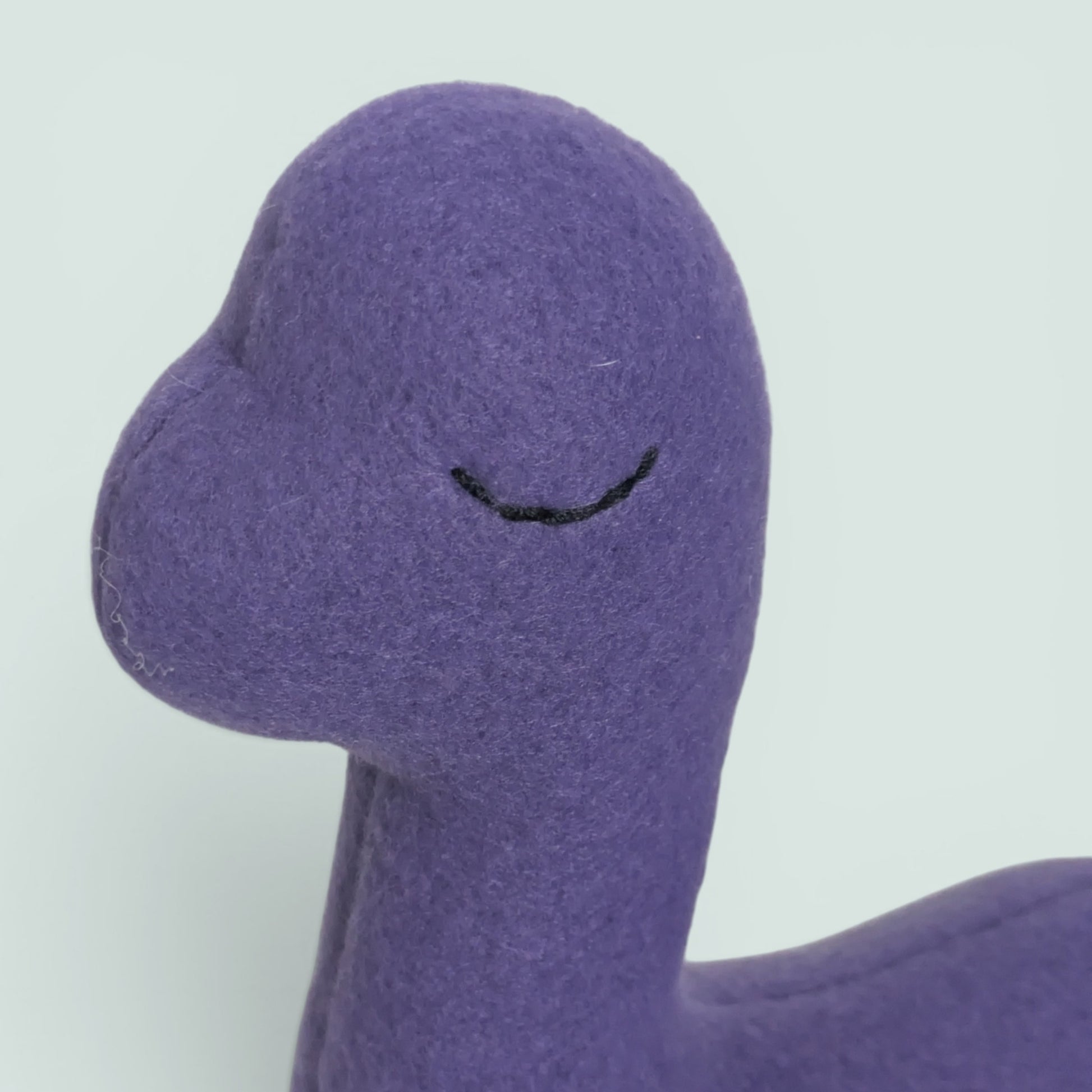 Close-up of a purple fleece plush toy with a simple black embroidered eye on a light gray background.