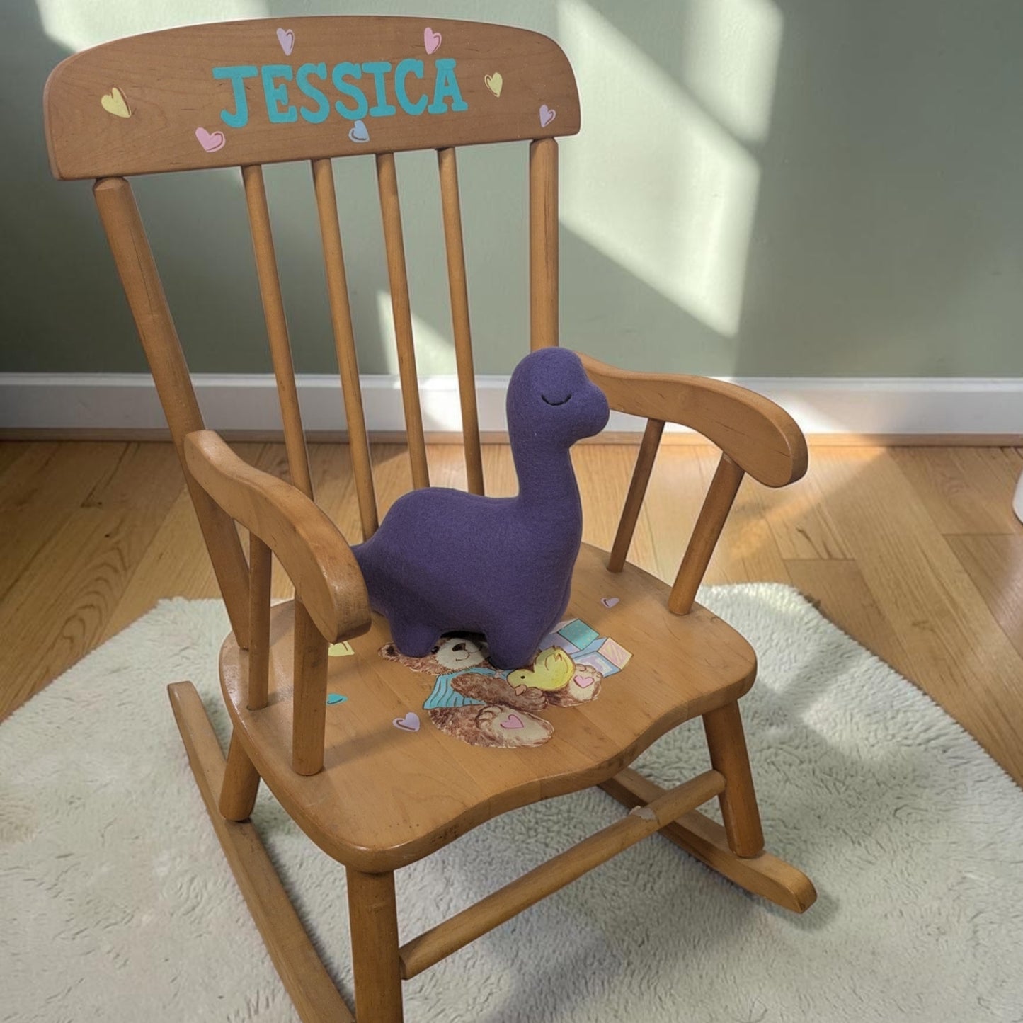 A wooden rocking chair with the name 'JESSICA' painted in blue on the backrest, decorated with small pastel hearts. A purple plush dinosaur toy sits on the chair, which is placed on a white rug over a wooden floor.