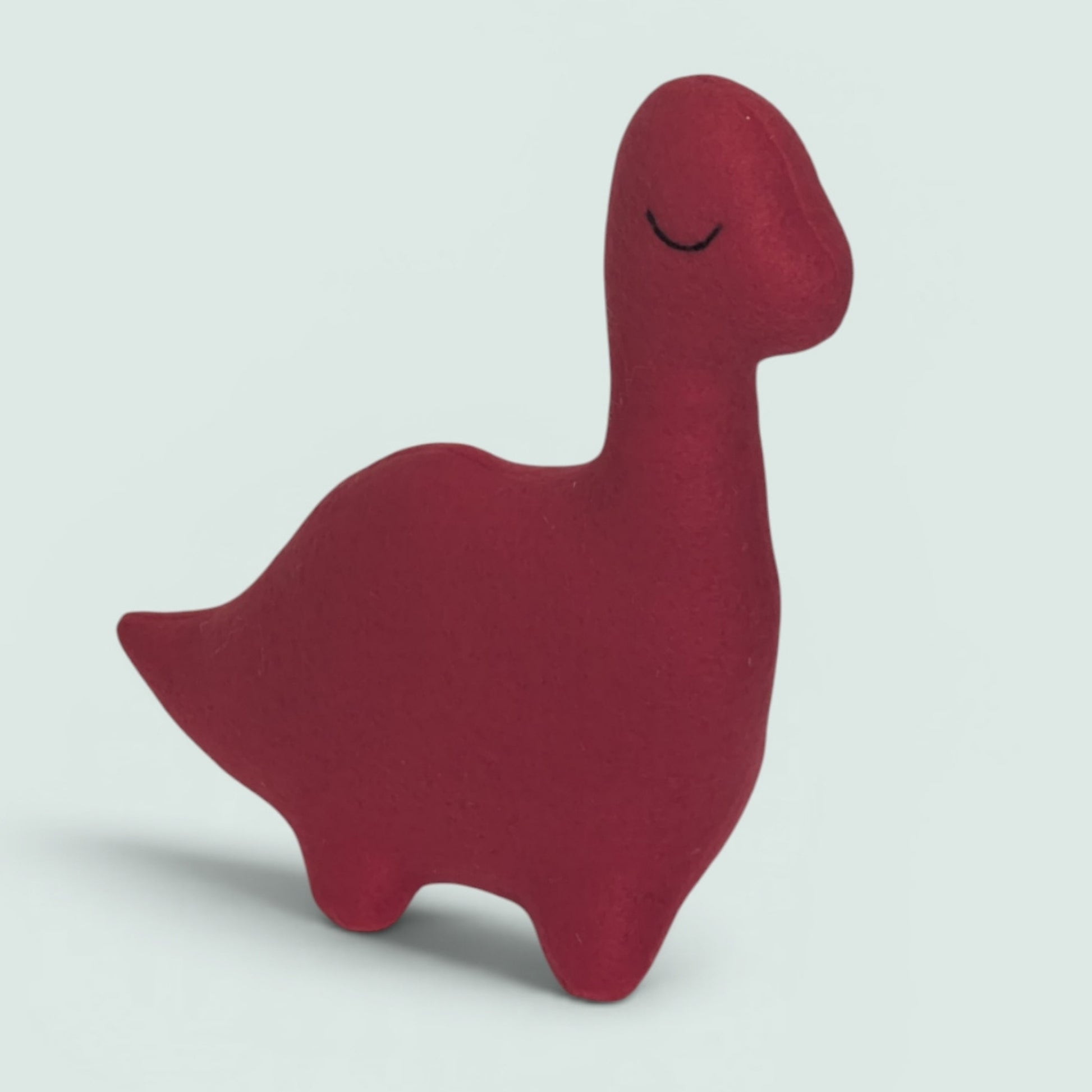 A small handmade plush dinosaur toy in red, featuring a simple embroidered closed eye on its head, set against a solid light gray background.