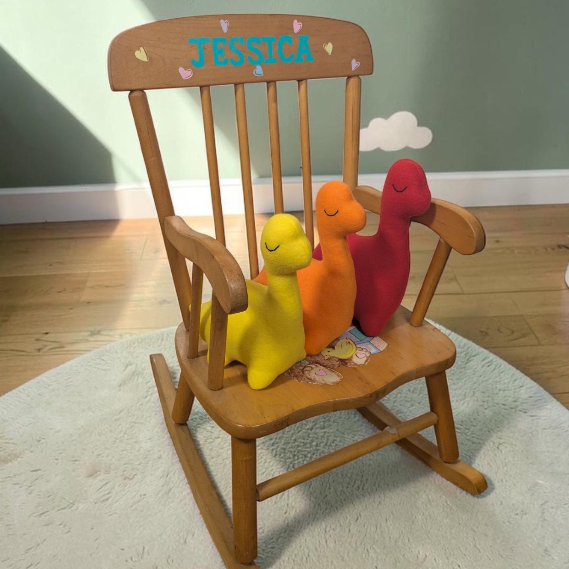 A wooden rocking chair with a personalized name 'JESSICA' on the backrest, styled with three small plush dinosaur toys in yellow, orange, and red on a white rug over a wooden floor.