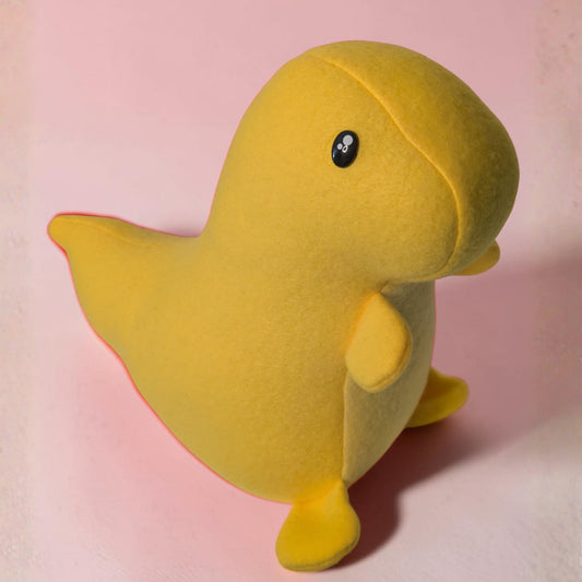 A yellow plush toy shaped like a cartoon dinosaur, standing on a soft pink background. The toy has a small black safety eye with white details.