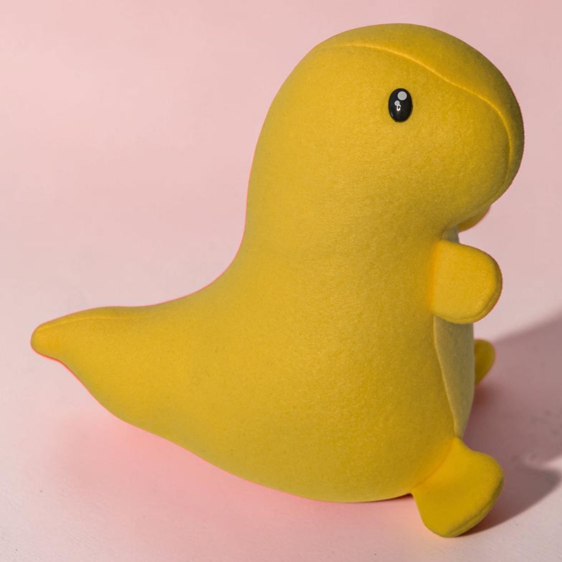 A bright yellow plush toy shaped like a dinosaur, photographed against a soft pink background.