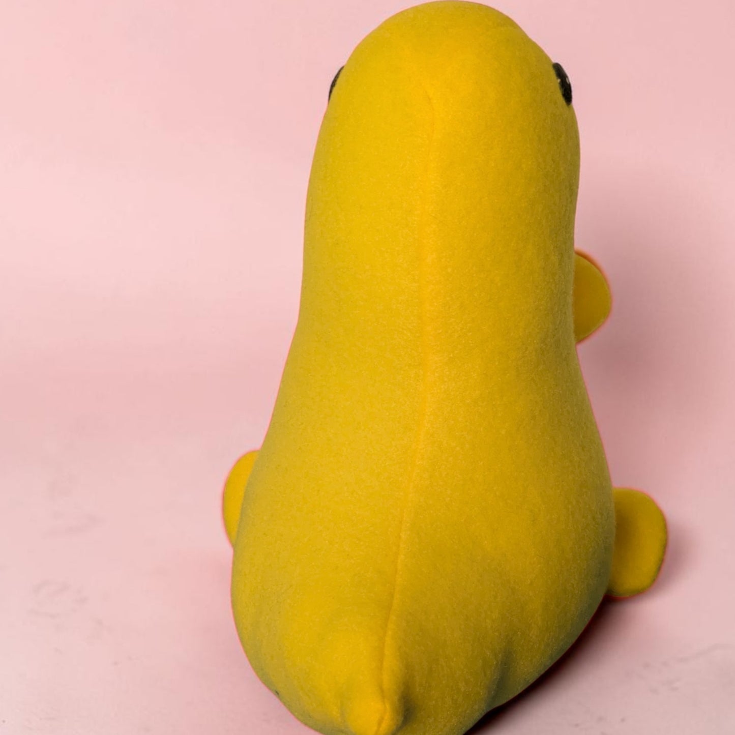 A bright yellow plush toy shaped like a dinosaur, photographed from the back against a soft pink background.