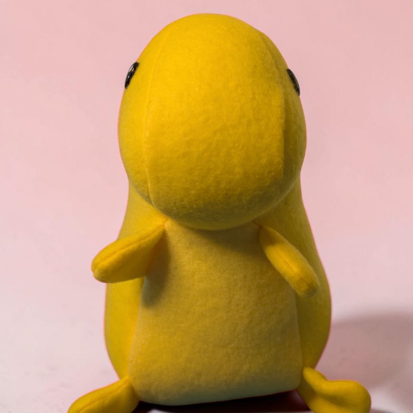 A yellow plush dinosaur toy with a smooth, rounded body, small black eyes, and tiny yellow limbs, photographed against a soft pink background.