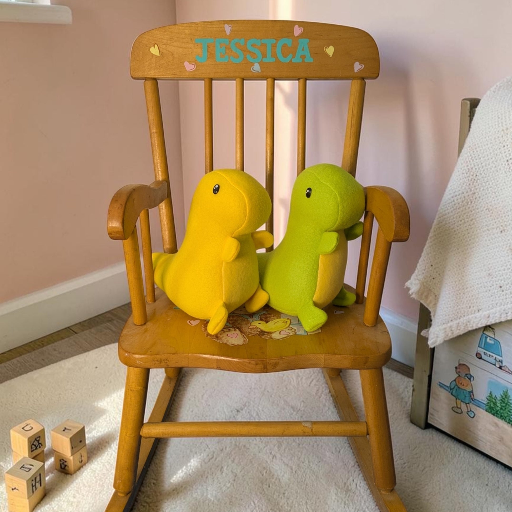 A wooden rocking chair with the name 'JESSICA' painted on the backrest, holding two plush dinosaur toys, one yellow and one green, in a child's room.