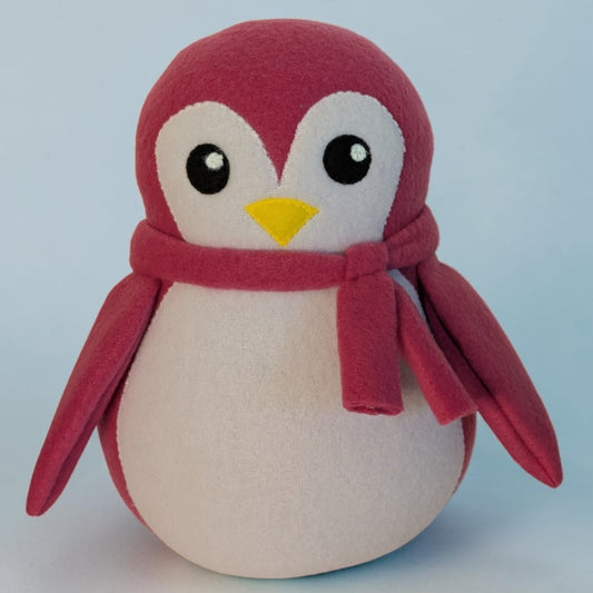 A plush penguin toy with a pink fleece body, white face and belly, black eyes, and a small yellow beak, wearing a matching pink scarf, photographed against a light blue background.