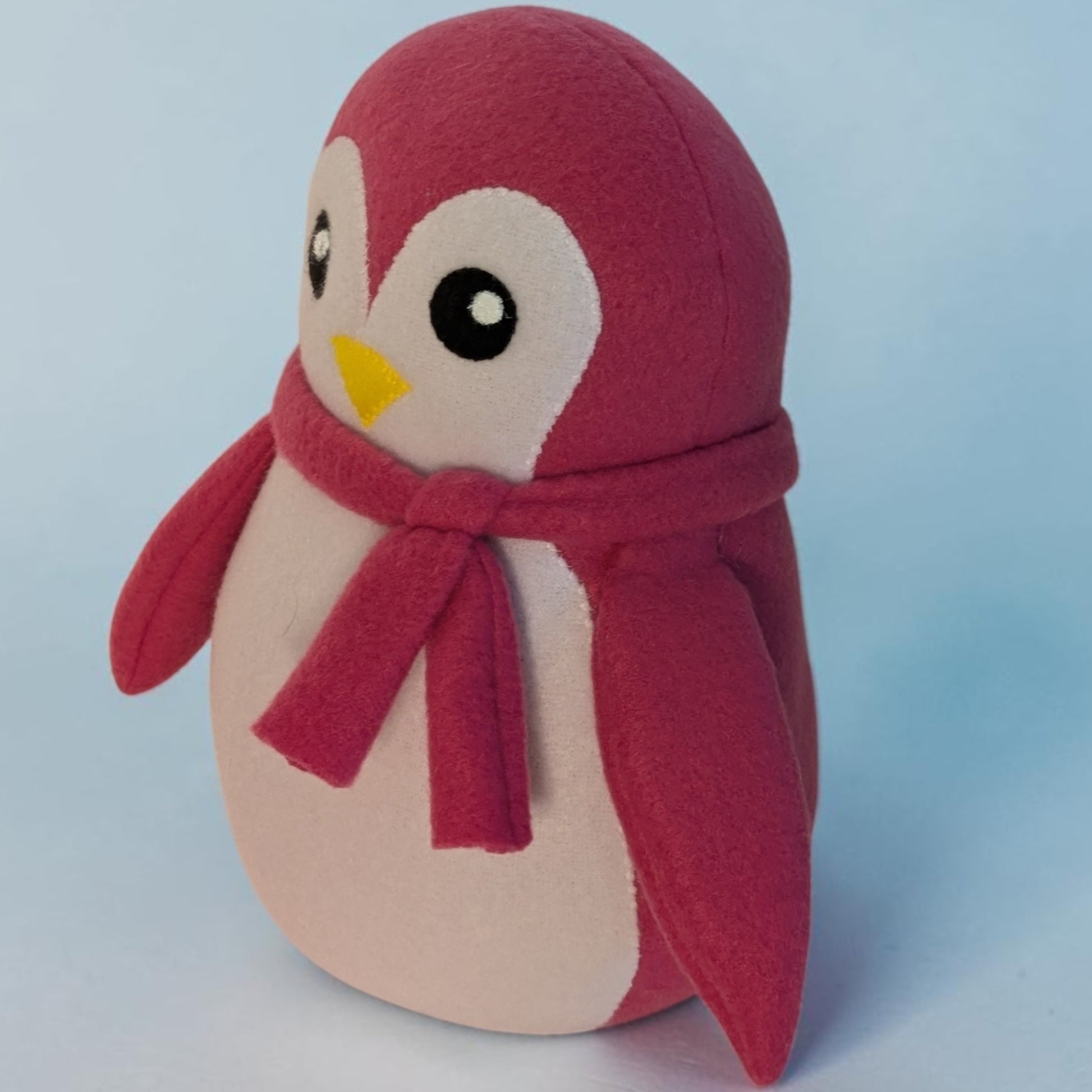 A plush penguin toy with a pink fleece body, a white belly, a matching pink scarf, and a small yellow beak, photographed against a soft light blue background.