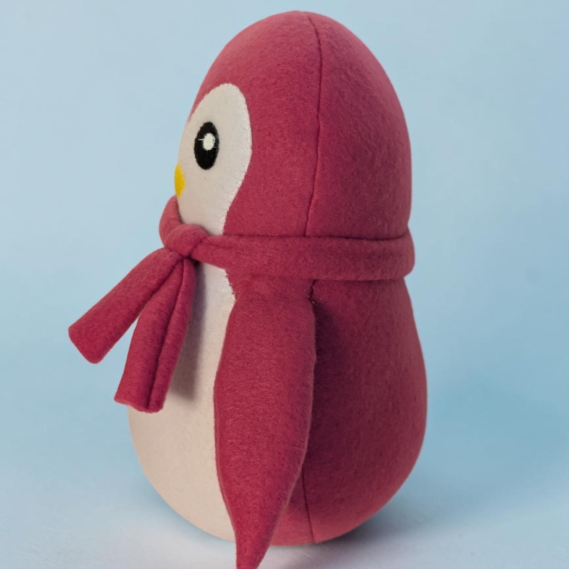 A plush penguin toy with a dark pink fleece body and scarf, a cream-colored belly and face, and a small yellow beak, set against a light blue background.