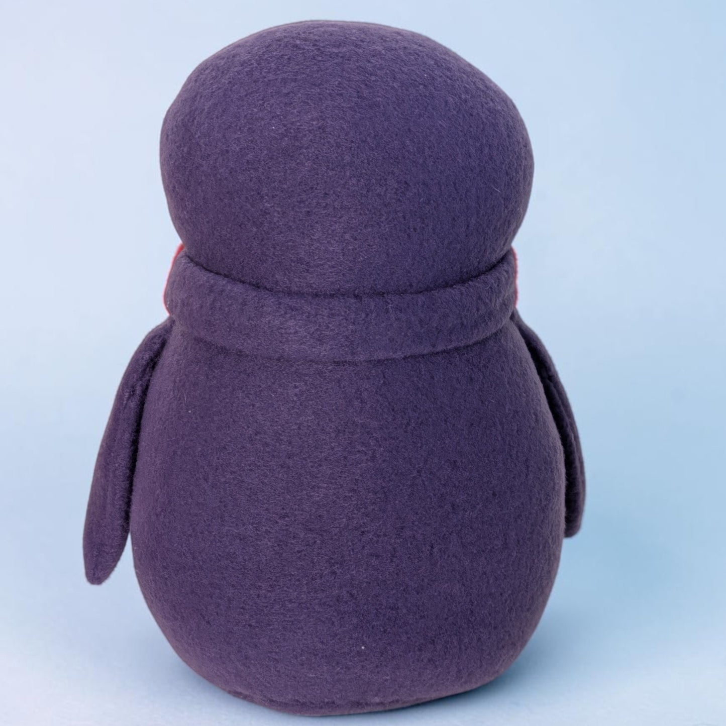 A purple fleece penguin stuffed animal photographed from the back against a light blue studio background.