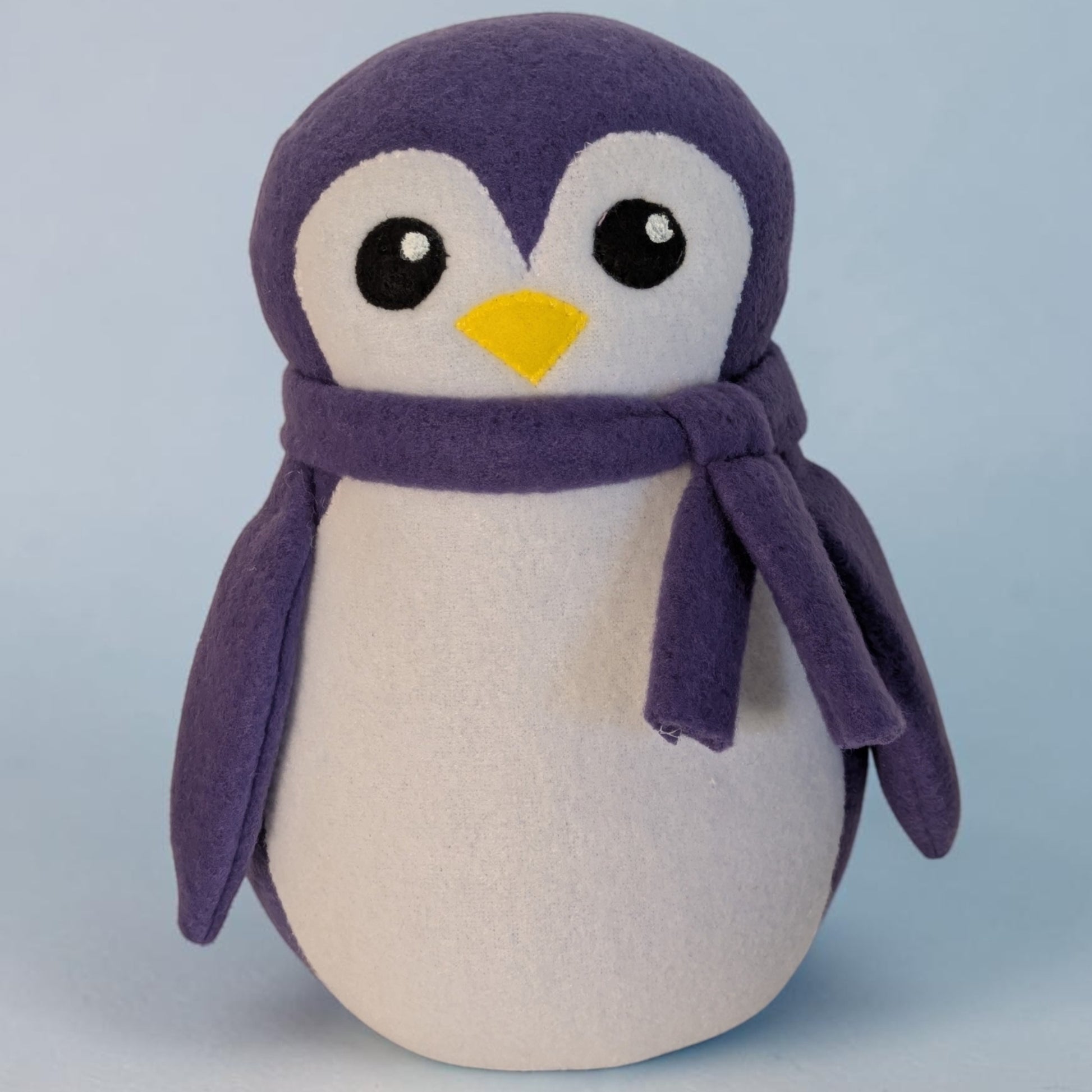 A plush penguin toy with a purple body and scarf, a white belly and face, and a small yellow beak, photographed against a soft light blue background.