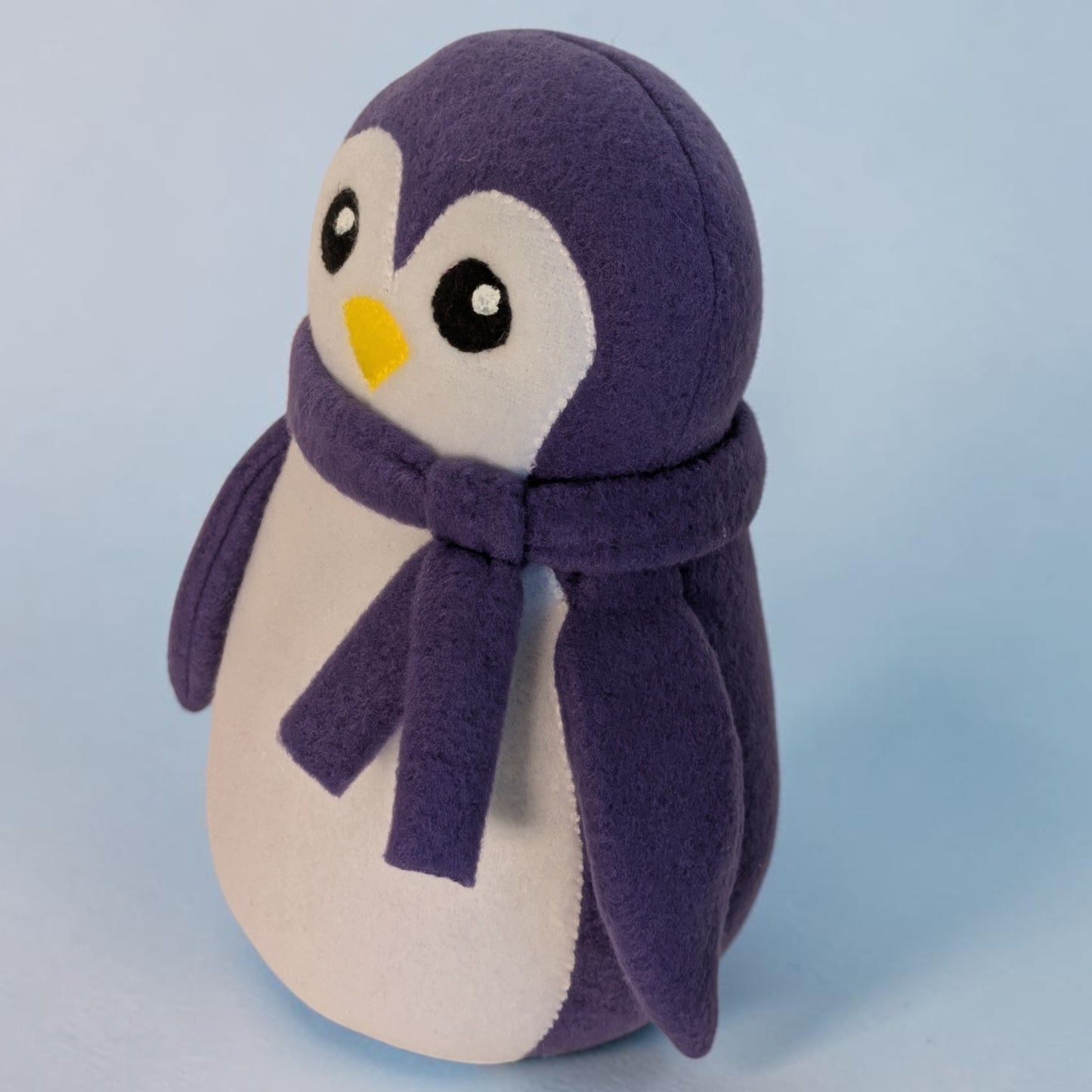 A plush penguin toy with a purple fleece body, white belly, matching purple scarf, and a small yellow beak, photographed against a soft light blue background.