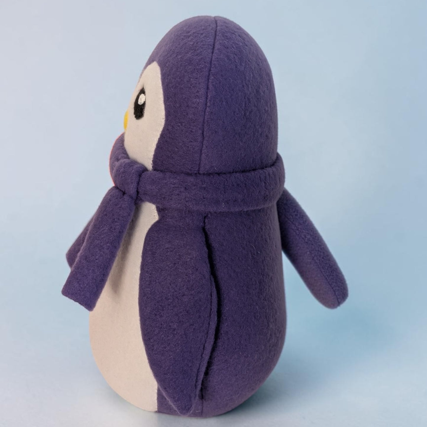 A plush penguin toy in purple and cream, wearing a matching purple scarf, photographed against a soft light blue background.