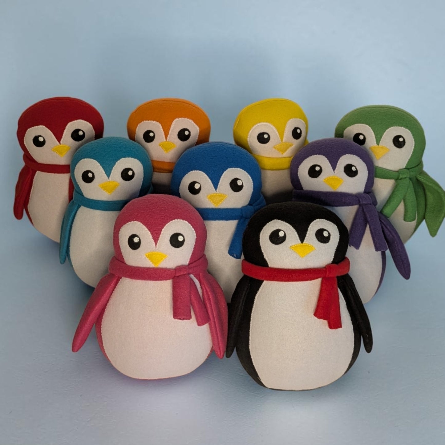 A set of nine soft plush penguin toys arranged together on a light blue background. Each penguin has a different color body and scarf. They are red, orange, yellow, teal, blue, purple, pink, and black, with a white belly and face,, black eyes, and a yellow beak.