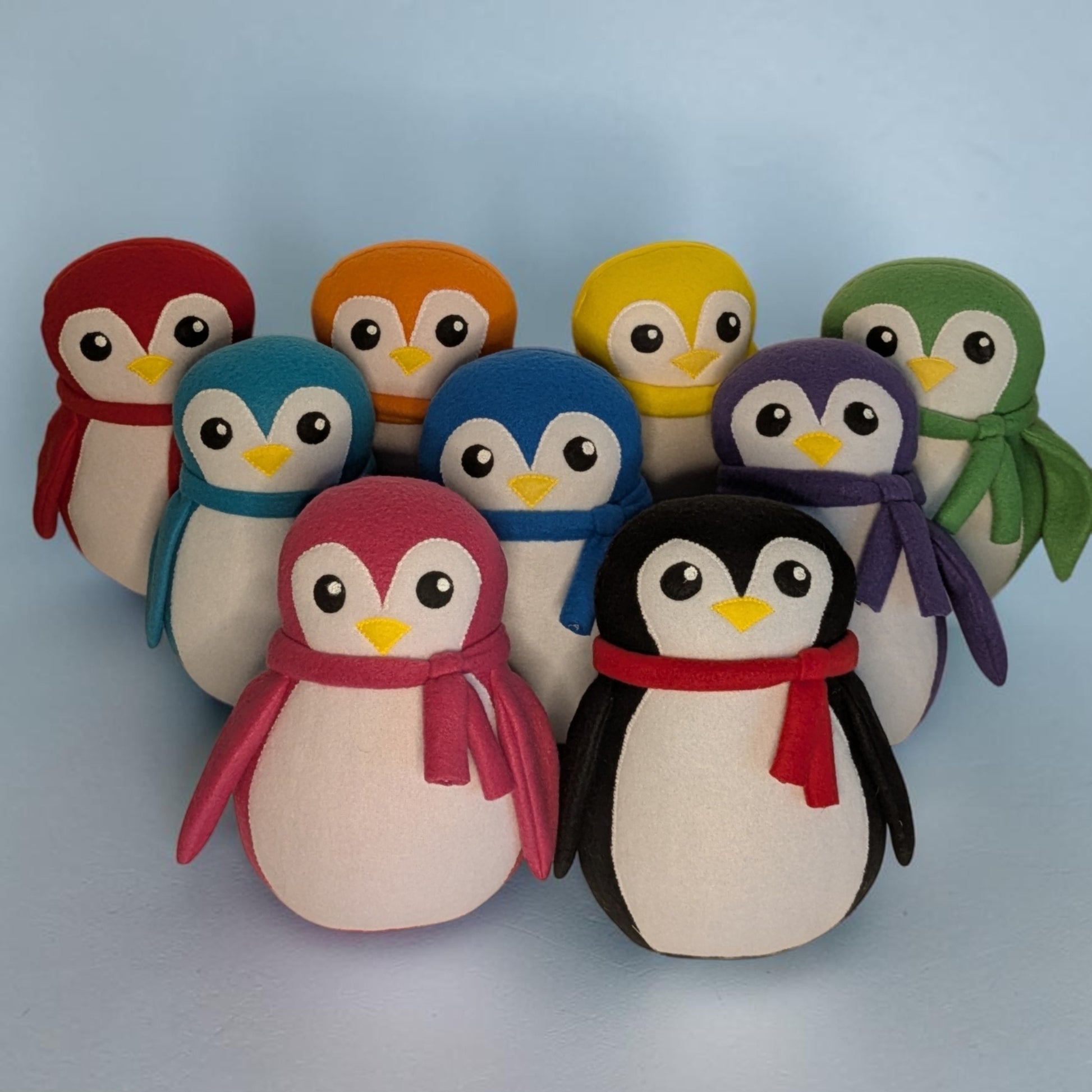 A set of nine soft plush penguin toys arranged together on a light blue background. Each penguin has a different color body and scarf. They are red, orange, yellow, teal, blue, purple, pink, and black, with a white belly and face,, black eyes, and a yellow beak.