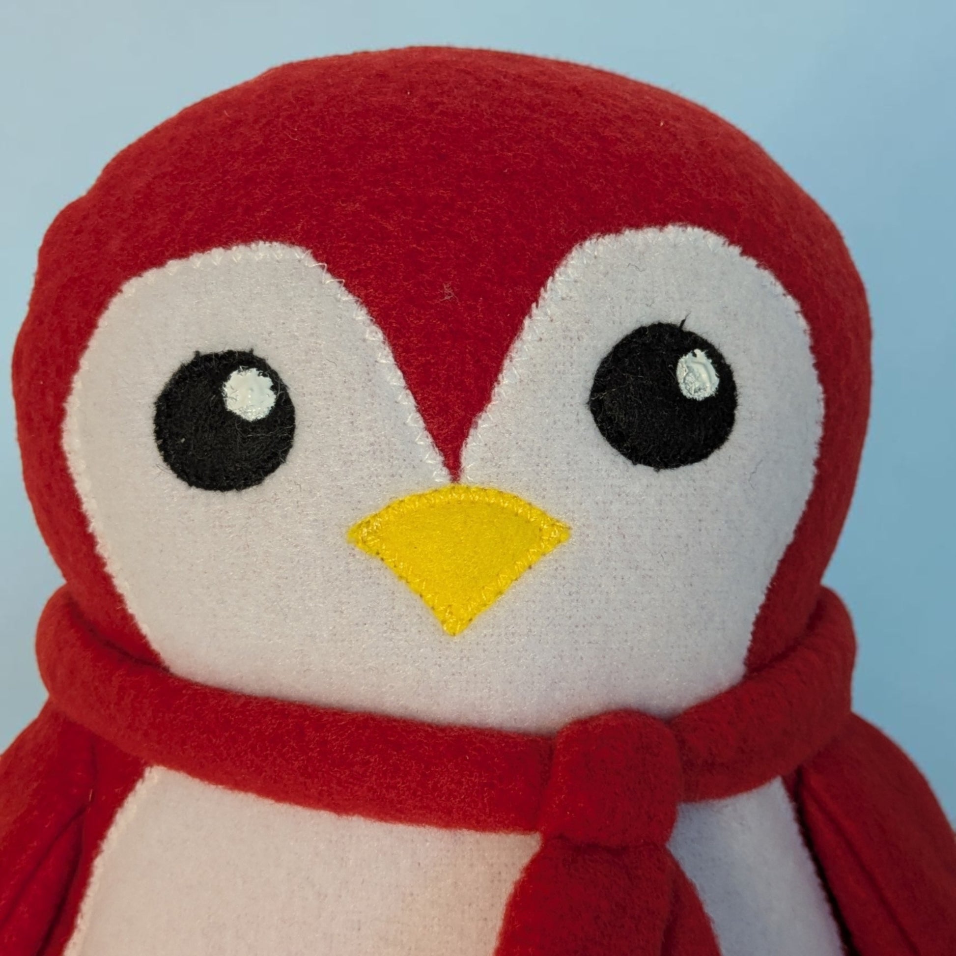 A close-up of a red and white plush penguin toy wearing a red scarf, with black felt eyes and a yellow beak, set against a light blue background.