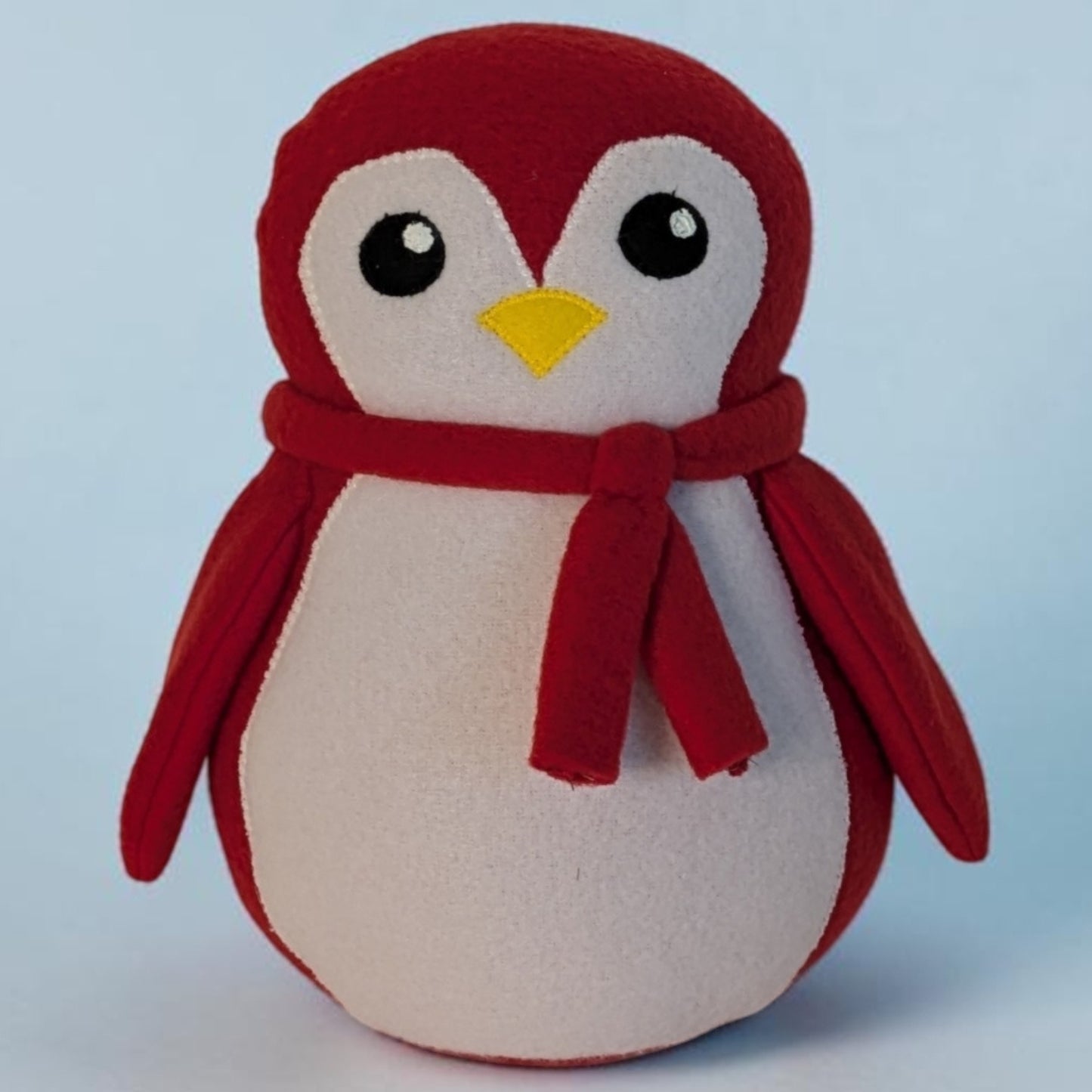 A plush penguin toy with a red body, white belly and face, yellow beak, and a red scarf, photographed against a light blue background.
