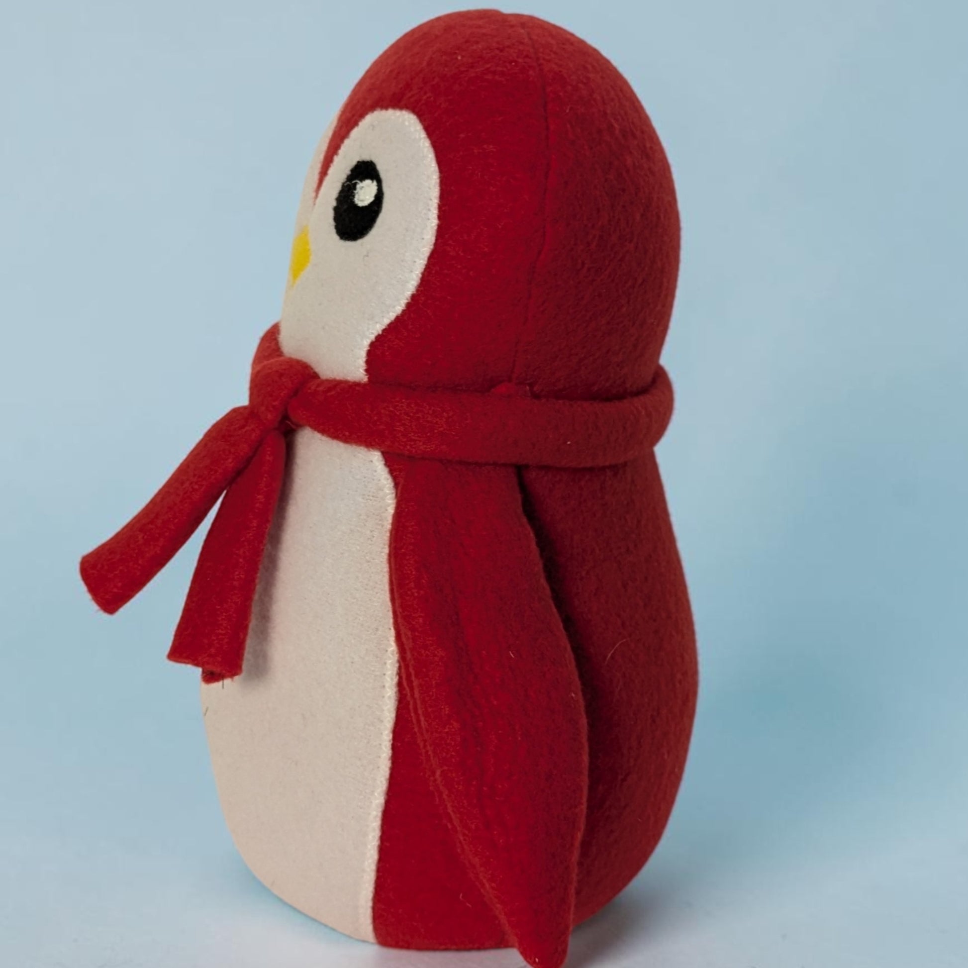 A red and white plush penguin toy wearing a matching red scarf, photographed against a light blue studio background.