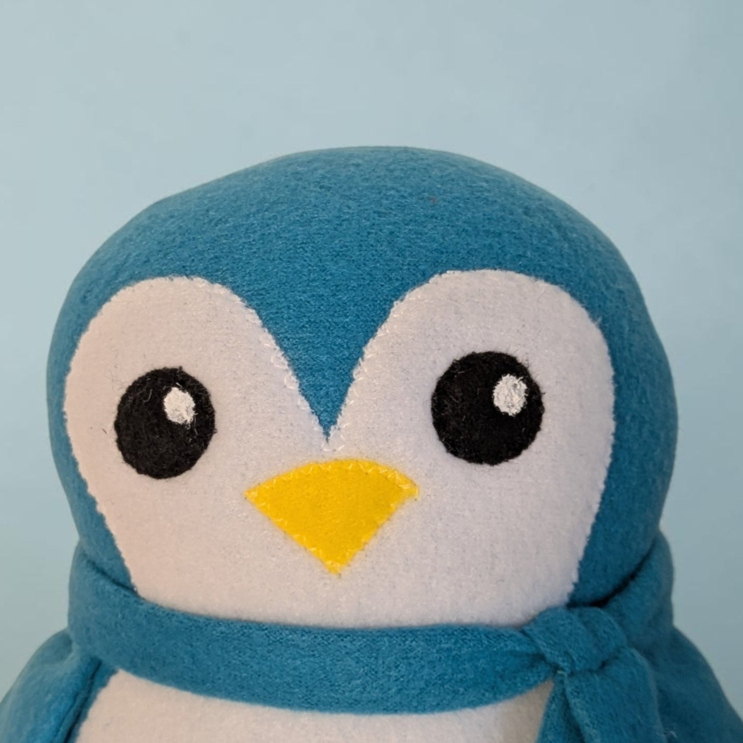 A close-up of a blue and white plush penguin toy wearing a matching blue scarf, set against a solid light blue background.