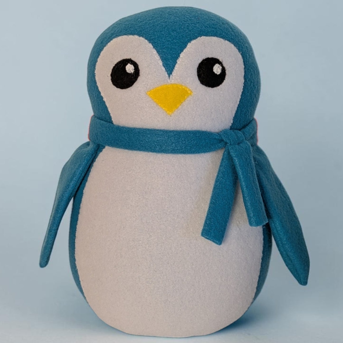 A plush penguin toy with a blue head and wings, a cream-colored belly, a yellow beak, and a blue scarf, photographed against a light blue background.