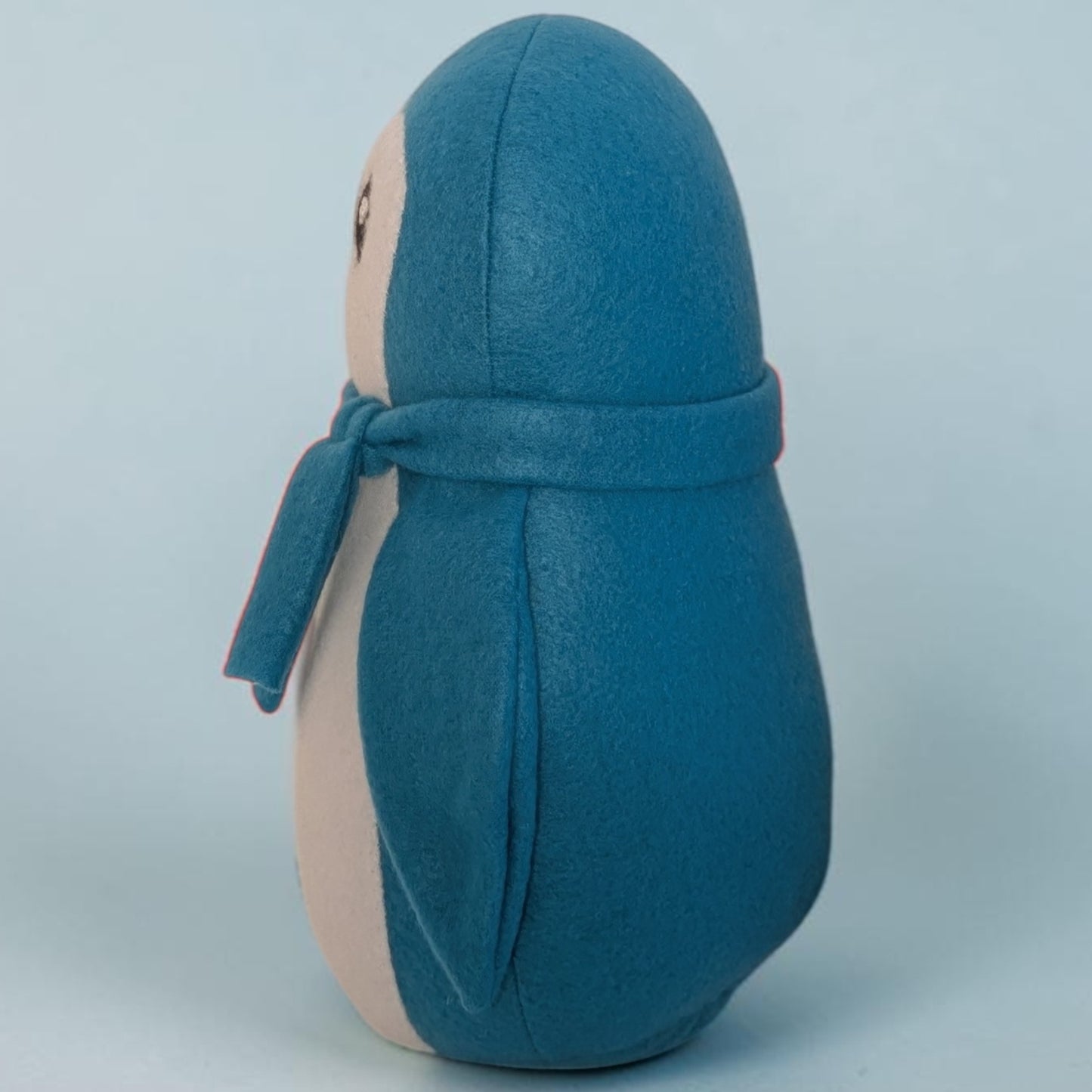 A small plush penguin toy shown from the side, with a blue body, white  belly, and a matching blue scarf, photographed against a light blue-gray background.
