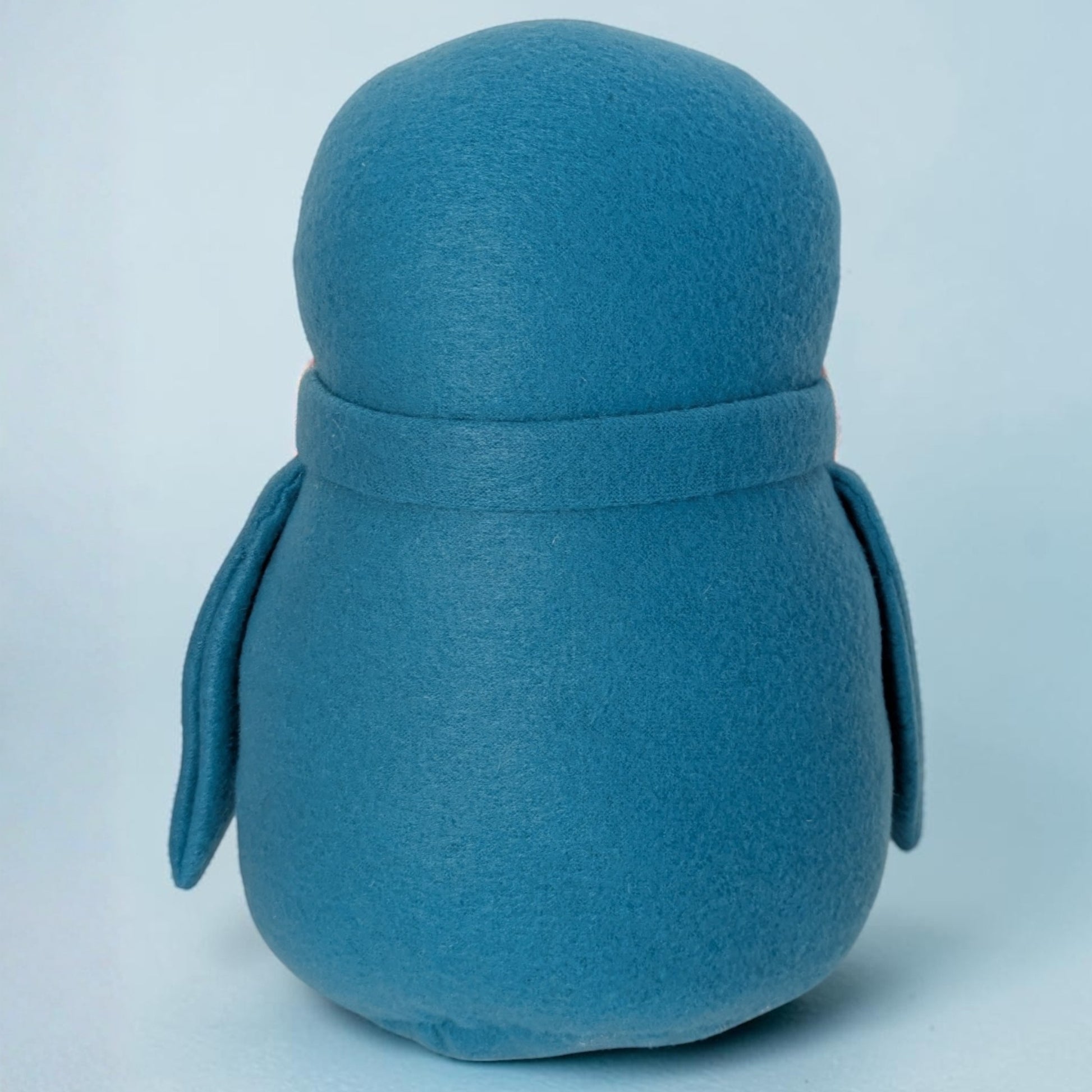 A blue fleece penguin stuffed animal photographed from the back against a light blue studio background.