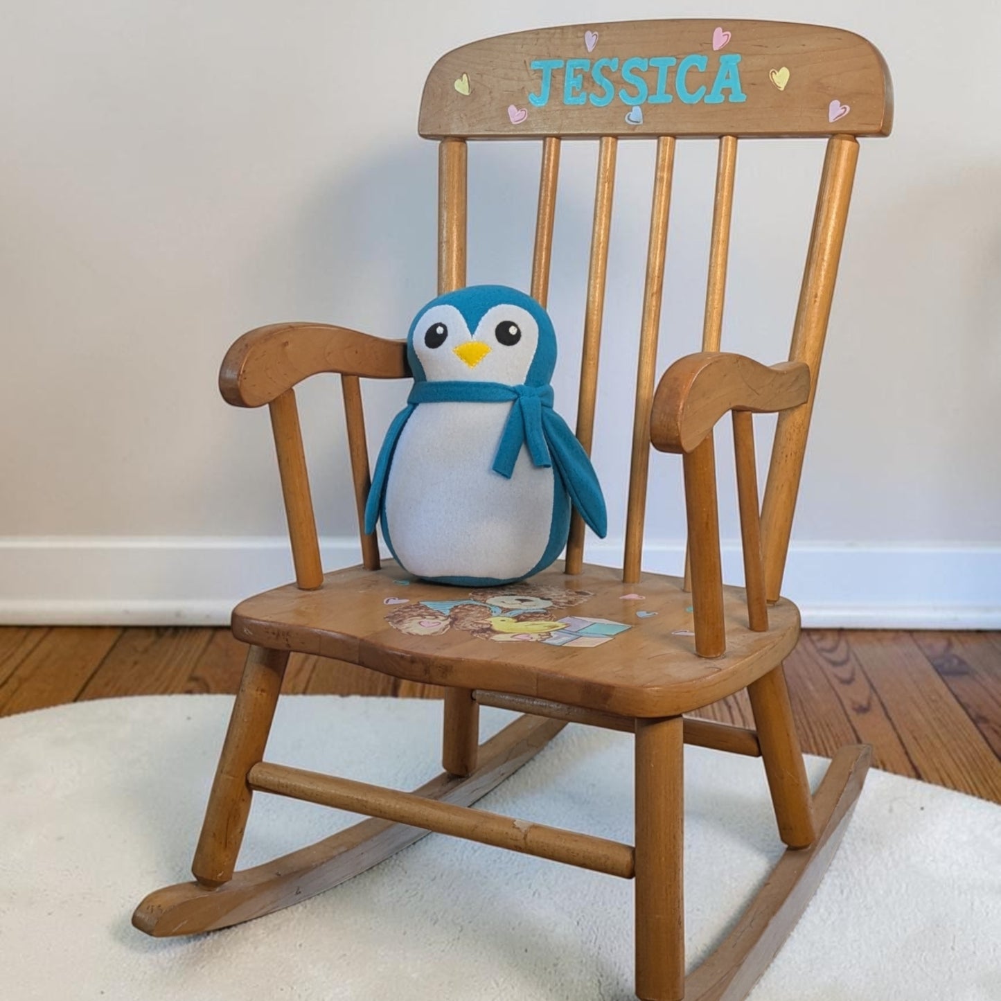 A wooden rocking chair with the name 'JESSICA' painted on the backrest and a plush penguin toy sitting on the seat, placed in a home setting on a white rug over a wood floor.